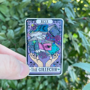 K&ouml;nnte beinhalten: Eine violette Tarotkarte mit holografischem Finish. Die Karte zeigt ein Paar H&auml;nde, die einen Stapel B&uuml;cher halten. Die Kartennummer ist XCIX und der Titel lautet "THE COLLECTOR".