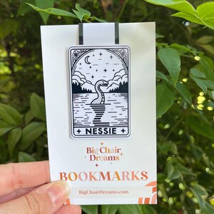 May include: A black and white Nessie bookmark featuring a lake scene with mountains, stars, and a crescent moon. The bookmark is held by a hand and has the text "NESSIE" and "Big Chair Dreams BOOKMARKS" printed on it.