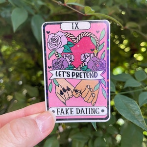 以下が含まれることがあります： IXと「Let's Pretend Fake Dating」の文字が書かれたピンクとパープルのタロットカード。カードには、ロープでできたハートの形と、その周りに花が飾られており、2つの手が互いに握り合っている。