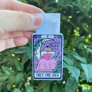 May include: A purple and green tarot card with a cartoon illustration of a bear sleeping in a bed with a heart-shaped pillow. The card reads "And there was... ONLY ONE BED" and has a keyhole symbol in the top right corner.
