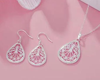 May include: A silver jewelry set featuring a pendant necklace and matching earrings. The teardrop-shaped pieces have an open filigree design with a delicate pattern. The set is displayed on a pink surface.