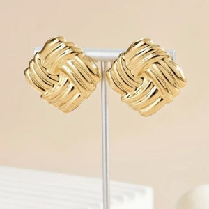 May include: A pair of gold-tone knot-shaped earrings. The earrings are made of a textured metal and have a square shape.
