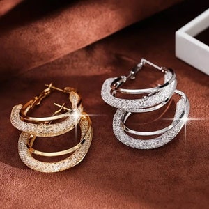 May include: A pair of hoop earrings, one gold and one silver, with a double hoop design. Both earrings are covered in small, sparkling rhinestones.