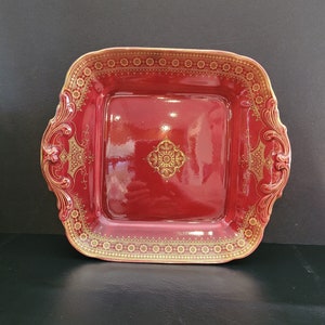 May include: A square, red ceramic serving dish with gold filigree detailing. The dish has a raised rim and ornate corner designs. The centre features a gold medallion. The overall design is symmetrical and elegant, suitable for serving appetizers or desserts.