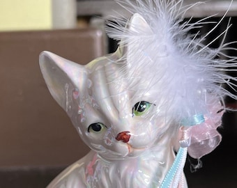 May include: A white porcelain figurine of a cat wearing a white feathered hat with a pink and blue ribbon. The cat has green eyes and a red nose.