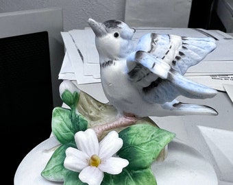 May include: A white ceramic trinket box with a blue bird perched on a branch with green leaves and a white flower. The bird has black eyes and a black beak.