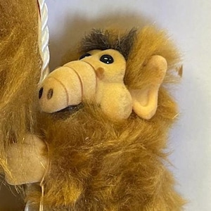 Puede incluir: Un peluche del personaje ALF, con hocico, orejas y pelaje de color beige. El juguete tiene una etiqueta roja con la palabra "ALF" en letras amarillas. El juguete está sujeto a una cuerda blanca.