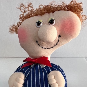 Könnte beinhalten: Handgemachte Puppe mit Stoffgesicht, rosigen Wangen und lockigem braunem Haar. Die Puppe trägt ein blau-weiß gestreiftes Outfit mit roter Fliege. Die Arme und Beine der Puppe sind ebenfalls aus Stoff.