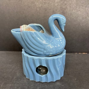 Pode incluir: Um aquecedor de óleo em forma de cisne de cerâmica azul claro. O cisne está sobre uma base correspondente com um design esculpido e uma pequena abertura. A parte de trás aberta do cisne contém um pequeno saco.
