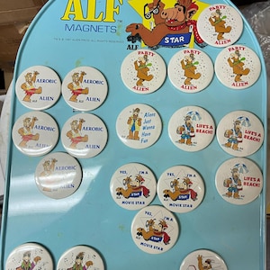 May include: A collection of ALF magnets on a light blue magnetic board. The magnets feature various images of the ALF character, with phrases like "Aerobic Alien", "Party Alien", and "Life's a Beach!" The board has the ALF logo.