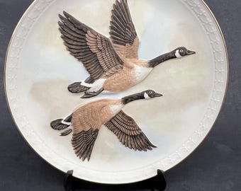 Op de afbeelding: Een witte porseleinen plaat met een gouden rand heeft een 3D-afbeelding van twee Canadese ganzen in vlucht. De ganzen zijn bruin en wit met zwarte vleugelpunten. De plaat staat op een zwarte standaard.