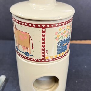 May include: A ceramic candle warmer with a white base and a removable top. The top features a colourful design with a cow, flowers, and a bucket. The base has a hole in the centre for a tea light candle. The warmer is marked with the copyright symbol and the letters "CGD".