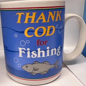 May include: A blue ceramic coffee mug with a white handle. The mug has a white rim and a red, white, and green stripe around the bottom. The mug has a cartoon fish and the text "THANK COD for Fishing" in yellow, white, and red.