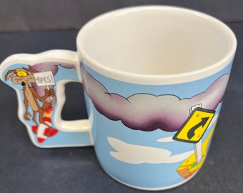May include: A white ceramic mug with a blue background and a cartoon coyote on the handle. The coyote is wearing a red rocket and holding a sign that says "Yipes!" The mug has a cartoon image of a road sign with a curved arrow on it.