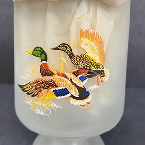Può includere: Un calice di vetro smerigliato con un disegno di due anatre in volo. Le anatre sono dipinte in tonalità di marrone, arancio, giallo, verde e blu.
