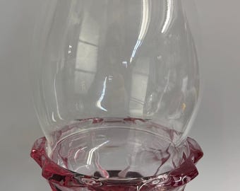 May include: A clear glass hurricane candle holder with a flared top and a ruby red base. The base has a decorative, scalloped edge. The candle holder is designed to hold a single candle.