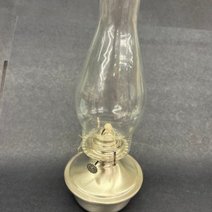 Könnte beinhalten: Eine silberne Öllampe mit einem klaren Glaszylinder. Die Lampe hat einen dekorativen Metallsockel und einen Dochtträger.