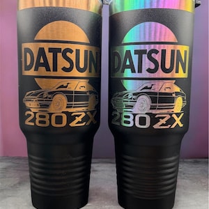 May include: Two matte black tumblers with clear lids, each featuring a laser-engraved graphic of a Datsun 280ZX car. The left tumbler has a metallic gold circular background, while the right tumbler displays a vibrant holographic circular design.