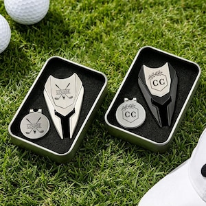May include: Two personalized golf divot tools and ball markers in metal tins, set on green grass. One set is silver with "LOUISE" and crossed golf clubs, the other is black and silver with "CC" and a laurel wreath. Golf balls are visible.