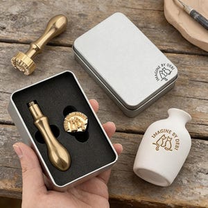 Könnte beinhalten: Ein Set aus goldfarbenen Wachssiegelstempeln und eine kleine weiße Vase mit dem Text "IMAGINE BY DEBI". Das Set enthält einen Stempel mit Griff, einen Stempelkopf und eine kleine Metallbox. Die Vase hat ein Katzenmotiv.