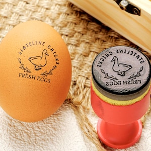 May include: An egg with a stamped design and a rubber stamp. The egg is light brown with the text "SIATELINE CHICKS FRESH EGGS" around a duck illustration. The stamp has the same design and is on a red egg cup.
