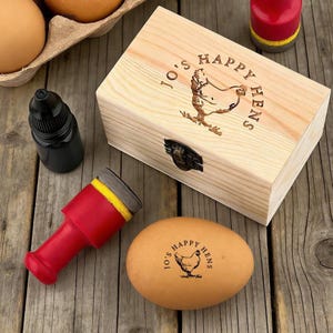 May include: A wooden box and an egg with the text "Jo's Happy Hens" and a hen illustration. A red and yellow stamp and a black ink bottle are also present. The box has a metal clasp.