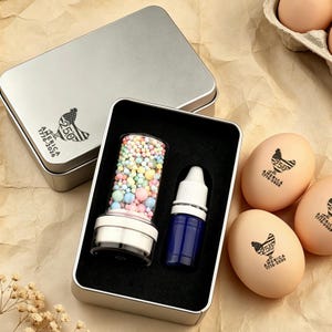 May include: A silver metal tin with a black interior holds a clear container filled with colorful spheres and a blue bottle with a white cap. The tin has a patriotic design with a chicken and the text "250 America 1776-2026".