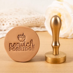 Könnte beinhalten: Ein runder, hellbrauner Wachssiegelstempel mit den Worten "norwich PORRIDGE" und einer Schüsselgrafik. Ein goldfarbener Metallgriff befindet sich neben dem Stempel. Der Stempel und der Griff befinden sich auf einer hellbraunen Oberfläche.