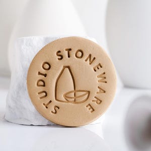 Peut inclure: Une pièce ronde en grès beige avec l'inscription "STUDIO STONEWARE" encerclant un motif central représentant une bouteille et un bol. La pièce est posée sur un fond blanc texturé avec des objets en céramique blancs flous.