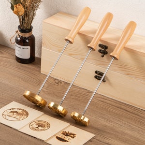 Può includere: Tre ferri da marcatura con manici in legno e teste in ottone, esposti con targhe in legno incise. I ferri hanno lunghi steli argentati. Le targhe presentano disegni circolari e floreali, con testo.