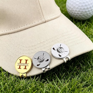 Könnte beinhalten: Drei personalisierte Golfballmarker mit Hutclips. Einer ist goldfarben mit der Gravur "Harmon", zwei sind silberfarben mit den Gravuren "Ashley" und "SZ POND". Ein Golfball und ein beiger Hut sind im Hintergrund.