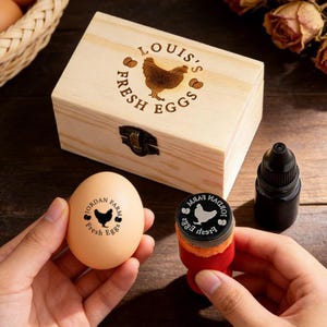 Può includere: Una scatola di legno con la scritta "LOUIS'S FRESH EGGS" e un'illustrazione di gallina. Un uovo è timbrato con "JORDAN FARM Fresh Eggs" e una gallina. Sono visibili anche un flacone di inchiostro nero e un timbro.