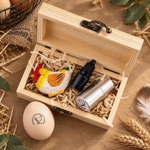 May include: A rectangular wooden box with a metal latch containing wood shavings, a small hand-painted wooden chicken figurine, a black plastic dropper bottle, and a silver metal cylinder. A single egg with the text Charlotte's Happy Hens sits nearby.
