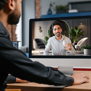 May include: A person in a black shirt is using a computer. The computer screen displays a video call with a man holding a glass of water. The background of the video call shows a modern interior with plants and chairs.