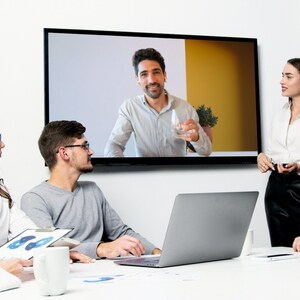 May include: A group of people are gathered around a table, looking at a screen showing a man on a video call. A woman is standing and talking to the group. There is a laptop computer on the table.