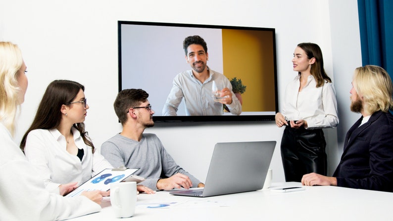 May include: A group of people are gathered around a table, looking at a screen showing a man on a video call. A woman is standing and talking to the group. There is a laptop computer on the table.