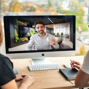 May include: Two people are sitting at a desk, looking at a computer screen. The person on the screen is holding a glass of water and smiling. The computer screen shows a modern office interior with a green wall.