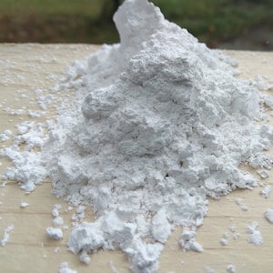 May include: A close-up shot of a pile of white powder on a light-colored wooden surface. The powder has a textured, granular appearance, with some larger clumps visible. The background is blurred, suggesting an outdoor setting.