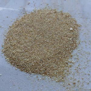 May include: A pile of light brown, granular material with small flecks of blue and white. The texture appears coarse and sandy, with a slightly uneven surface. The material is spread out on a white surface.