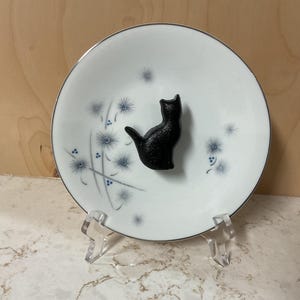 May include: A white ceramic plate with a silver rim and a black cat figurine. The plate has a floral design in blue and is displayed on a clear acrylic stand. The cat is in a sitting position.