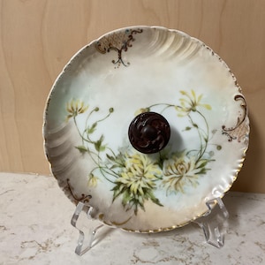 May include: Antique porcelain plate with a floral design featuring yellow flowers and green leaves. The plate has a scalloped edge with gold trim and a dark brown knob in the center. The plate is displayed on a clear acrylic stand.