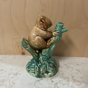 May include: A ceramic figurine of a koala bear perched on a green tree branch. The koala is brown with a glossy finish, while the branch and base are a contrasting shade of green. The figurine has a detailed, handcrafted appearance.