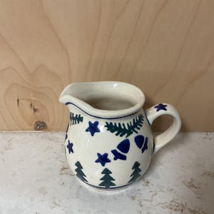 May include: A small, white ceramic creamer with a rounded body and a handle. The creamer is decorated with blue stars, fir trees, and other shapes. A blue line trims the rim and handle.