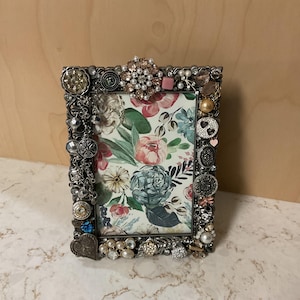May include: A rectangular picture frame with a floral print and embellished with various decorative buttons, pearls, and rhinestones. The frame has a silver finish and features a variety of shapes and sizes of embellishments.