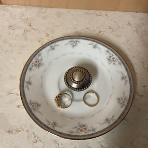 May include: A white porcelain ring dish with a floral pattern and gold trim. The dish holds a gold ring, a gold band, and a decorative metal knob. The dish is a decorative item for storing jewelry.