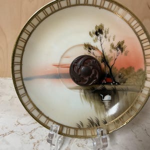 May include: A decorative antique plate with a hand-painted landscape scene featuring a sunset, trees, and a swan. The plate has a gold rim and a dark brown, ornate knob in the center. The plate is displayed on a clear stand.