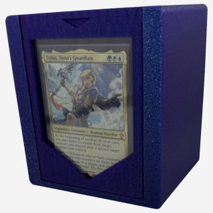 May include: A dark blue card storage box with a clear window displaying a Magic: The Gathering card. The card features an illustration of a blonde-haired character. The box has a textured, glittery finish.