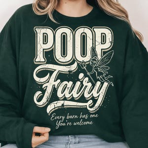Könnte beinhalten: Dunkelgrünes Sweatshirt mit den Worten "POOP Fairy" in abgenutzten weißen Buchstaben. Rechts vom Wort "Fairy" befindet sich eine Cartoon-Fee mit einer Mistgabel. Der Text "Every barn has one You're welcome" steht darunter.