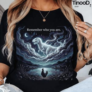 Puede incluir: Camiseta negra con una ilustración surrealista. Un dinosaurio emerge de las nubes bajo una luna creciente. El diseño incluye un lago, una gallina y el texto "Remember who you are."