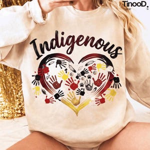 May include: Beige sweatshirt with the word "Indigenous" in black script above a heart-shaped design. The heart is filled with handprints in red, yellow, and black, along with feathers and paw prints. The sweatshirt has long sleeves.
